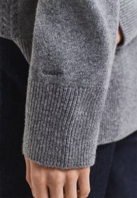 Manica di un maglione grigio in lana con polsino a coste, caratterizzata da una texture liscia e un sottile dettaglio di cucitura, indossata su uno sfondo scuro.