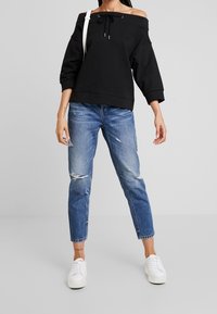 Felpa nera scollata sulle spalle con cordino, abbinata a jeans chiari strappati e sneakers bianche. Design semplice e casual.