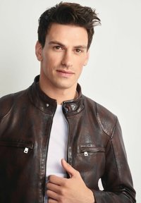 Młody mężczyzna z ciemnymi włosami, ubrany w brązową skórzaną kurtkę na białej koszuli, trzymający kołnierz kurtki na jasnym tle.