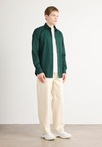 Langarm, donkergroene overhemd met zwarte knopen, gedragen over een witte T-shirt en gecombineerd met crèmekleurige wijde broek.