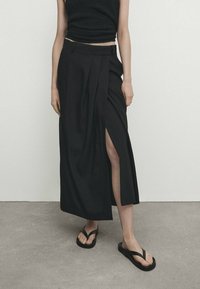 Jupe maxi plissée noire avec une fente sur le côté, en tissu léger. Associée à un débardeur noir et des tongs noires. Design simple.