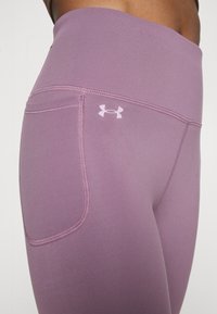 Fioletowe dopasowane legginsy z wysokim stanem, z małą kieszenią i kontrastowymi przeszyciami. Logo Under Armour jest wyświetlane w jasnofioletowym kolorze.