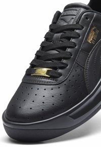 Sort læder sneakers med perforeret detaljer, flade sorte snørebånd, guld logo accenter og en afrundet gummisål.