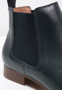 Stivale in pelle nera alla caviglia con pannello laterale elasticizzato e tacco in legno marrone basso, che mostra una texture liscia e cuciture dettagliate.