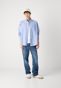 Blau-weiß gestreifte Button-Up-Shirts, darunter ein weißes T-Shirt, blaue Jeans, weiße Sneakers mit Gummisohle und eine beige gestrickte Mütze.