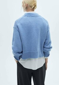 Blauer Strickpullover mit einem verkürzten Design, geripptem Saum und hochgeschlagenen Ärmeln. Über einem weißen, mehrlagigen Hemd getragen und mit dunklen Hosen kombiniert.