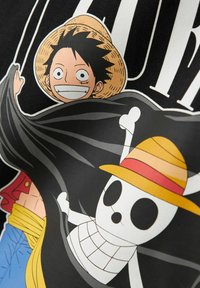 Lächelnder Anime-Charakter mit schwarzen Haaren und einem Strohhut, der hinter einer schwarzen Piratenflagge steht, auf der ein weißer Schädel mit passendem Strohhut abgebildet ist.