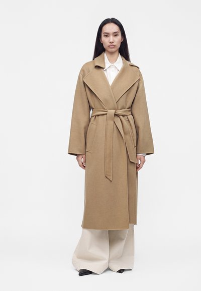 Femme portant un long manteau camel avec une ceinture sur une chemise blanche et un pantalon large beige, debout devant un fond clair uni.