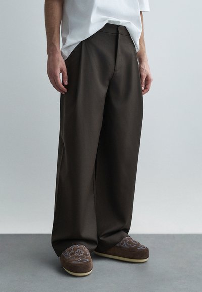 Pantalon à jambes larges en tissu marron foncé à texture lisse, avec un devant épuré et des poches. Associé à des mules brodées marron.