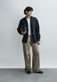 Camisola preta de manga comprida por cima de uma t-shirt branca, combinada com calças largas bege e ténis pretos, segurando uma mala preta.