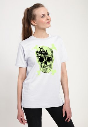 Žena s dlouhým culíkem, která má na sobě bílý tričko s potiskem zelené lebky a zkřížených kostí, doplněné černým graffiti textem.