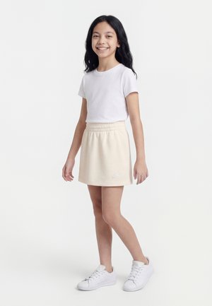 Fille souriante aux longs cheveux noirs, portant un t-shirt blanc, une jupe beige Nike et des baskets blanches, debout devant un fond blanc uni.