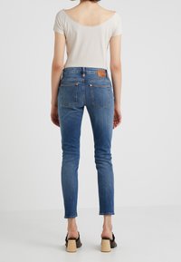 Siniset kapeat farkut korkealla vyötäröllä, joissa on ruskea nahkapatja takana, kontrastivirkkaukset ja klassiset takataskut.