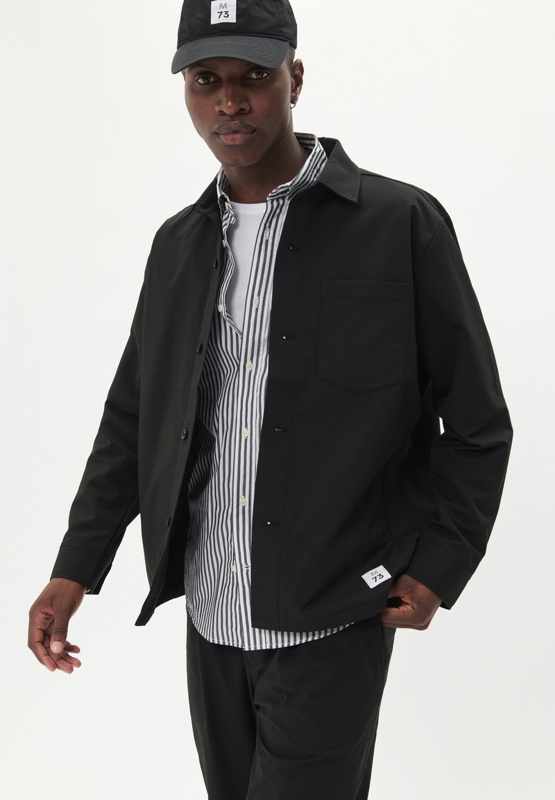 Czarna kurtka na guziki o gładkiej fakturze, z jedną kieszenią na piersi, noszona na białej koszuli w paski z kołnierzykiem, zestawiona z czapką w kolorze czarnym.