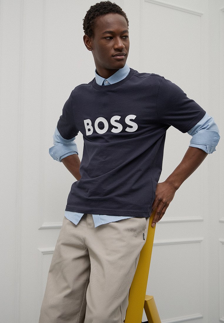 T-shirt en coton bleu marine avec le logo "BOSS" blanc, col rond, manches courtes, superposé sur une chemise à col bleu clair, assortie avec un pantalon beige.