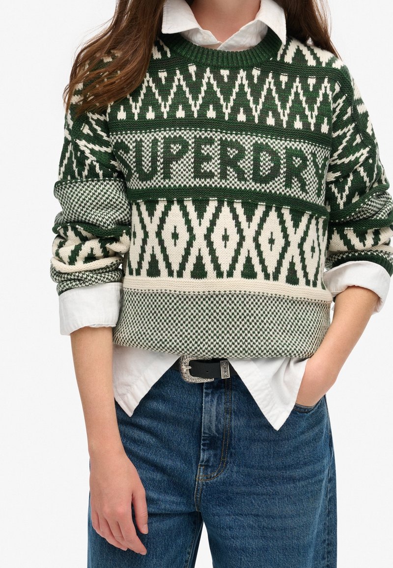 Personne portant un pull en tricot à motifs verts et blancs avec le texte « SUPERDRY », une chemise blanche en dessous, et un jean bleu avec une ceinture.