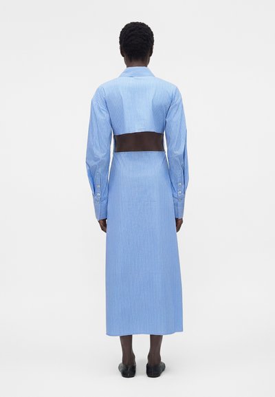 Άτομο με κοντά κατσαρά μαλλιά φοράει ένα μακρύ μπλε και άσπρο ριγέ φόρεμα με μεγάλο άνοιγμα στην πλάτη, στέκεται σε λευκό φόντο.