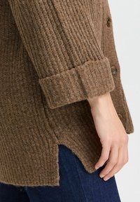 Brauner gerippter Wollcardigan mit seitlichem Schlitz, ausgestattet mit großen braunen Knöpfen und umgeschlagenem Ärmelsaum. Der Stoff hat ein strukturiertes, weiches Erscheinungsbild.