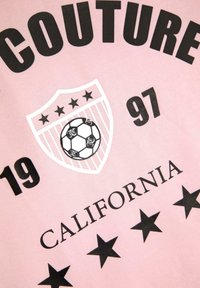 Růžové tričko s výrazným černým textem "COUTURE" a "CALIFORNIA", designem ve tvaru štítu se fotbalovým míčem a pěti černými hvězdami.