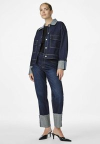 Farkkutakki, jossa on vaaleansininen kaulus ja hihansuut, yhdistettynä tummasinisiin farkkuihin, joissa on revenneet helmanteet. Mustat kärkitossut.