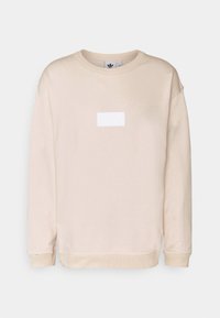 Beige sweatshirt with a crew neckline, ribbed cuffs, and hem. Features a rectangular white logo patch on the front. Soft fabric texture.