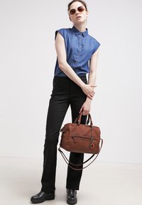 Brun lærveske med sømdetaljer, holdt i én hånd. Modell iført en denimskjorte og svarte bukser, med solbriller og svarte støvler.