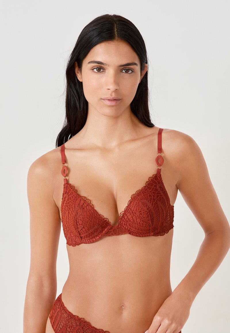 Bralette en dentelle bordeaux avec une forme triangulaire, des bords festonnés et quincaillerie métallique décorative sur les bretelles. Motif floral subtil sur l'ensemble.