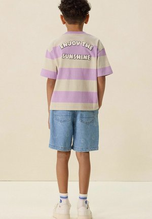 Enfant portant un t-shirt rayé violet et beige avec le texte « Profite du soleil », un short en jean bleu avec des visages souriants, des chaussettes blanches et des baskets.