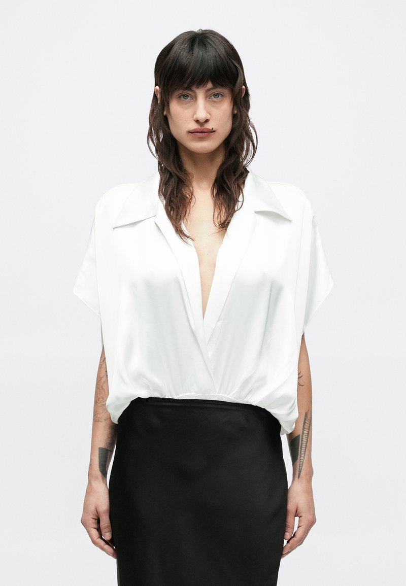 Bijela bluza od satena s dubokim V-izrezom i kratkim rukavima, uparena s visokim crnim suknjom. Glatka tekstura, krojena kroja, minimalistički stil.