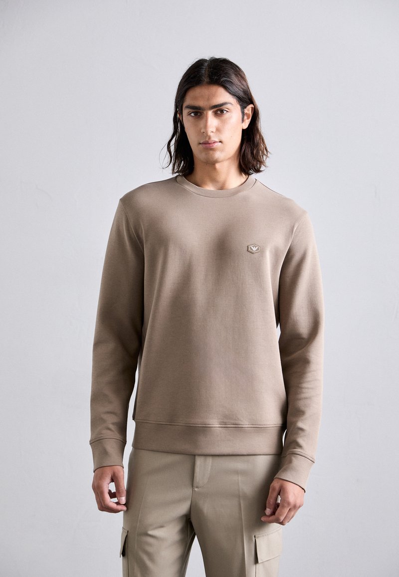Brown sweatshirt with a crew neck, long sleeves, and ribbed cuffs. Features a small, hexagonal logo on the chest. Soft texture and fitted design.