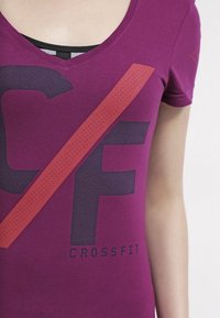 Detailní záběr na osobu ve fialovém tričku s výstřihem do V, na kterém je velký potisk "CF" a "CrossFit".