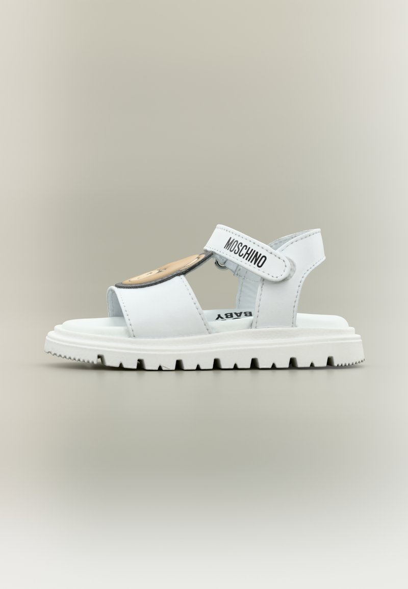 Λευκό σανδάλι για νήπιο με λουράκι Velcro που φέρει την επιγραφή "Moschino", ανθεκτική σόλα και λεπτομέρεια με πρόσωπο αρκουδάκι στο μπροστινό λουράκι.