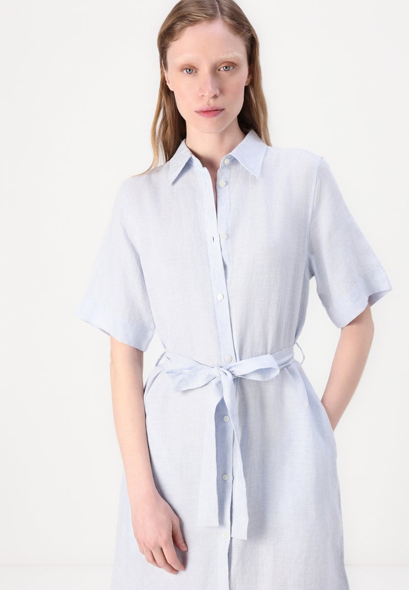 Helles blaues Kurzarmkleid mit einer Knopfleiste vorne und einem Bindegürtel in der Taille. Aus einem weichen, strukturierten Stoff gefertigt. Figurbetonter Schnitt mit Kragen.