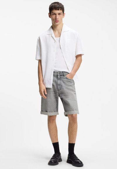 Jeune homme portant une chemise blanche à manches courtes boutonnée par-dessus un débardeur blanc, un short en jean gris retroussé, des chaussettes noires et des chaussures noires, debout.