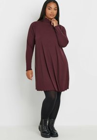 Lila ribbad klänning med hög halsringning och långa ärmar, som har en utställd silhuett och vågig nederkant. Bärs med svarta strumpbyxor och stövlar.