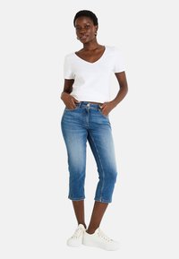 Camiseta blanca de cuello en V combinada con capris de mezclilla azules que tienen una cintura ajustada y un ligero desvanecimiento. La modelo lleva zapatillas blancas.