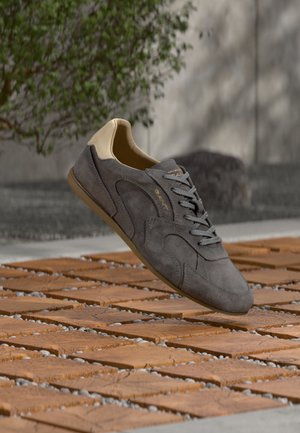Szary zamszowy but sportowy z beżowym paskiem na pięcie i gumową podeszwą, unoszący się nad drewnianą płytką i żwirową powierzchnią na zewnątrz.