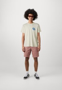 Light beige cotton t-shirt with blue logo on chest, paired with pink shorts. Model wears black and white sneakers with white socks. Red sunglasses.