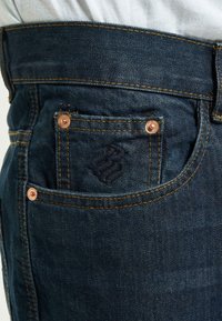 Jeans de mezclilla azul oscuro con herrajes en tono cobre, un pequeño logo bordado en el bolsillo trasero y detalles de costura en naranja contrastante.