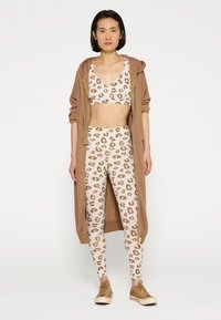 Bralette e leggings in stampa leopardo beige con macchie marroni, abbinati a una giacca con cappuccio marrone. Sneakers marroni completano il look.