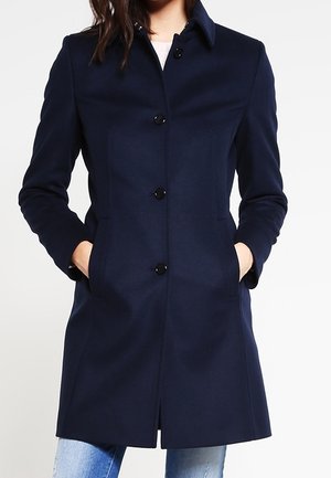 Frau trägt einen taillierten dunkelblauen, knöpfbaren Mantel mit Seitentaschen, kombiniert mit hellblauen Jeans, vor einfarbigem Hintergrund.