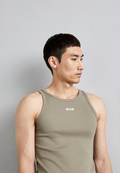 Tricou fără mâneci khaki cu textură cu striații, având decolteu tip crew și logo-ul alb "MSGM" în centru. Model cu părul scurt pe un fundal gri.