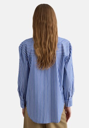 Donna con capelli lunghi e ondulati che indossa una camicia a righe verticali blu e bianche con bottoni, vista di spalle su uno sfondo neutro.