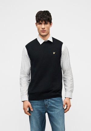 Man wearing black sleeveless sweater over white striped shirt and blue jeans, standing against plain light background.