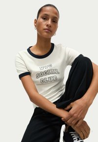 Katoenen t-shirt in off-white met een marineblauwe ribgebreide kraag, voorzien van "NYC SOCIAL CLUB" in dikke letters. Donkere broek met sneakers.
