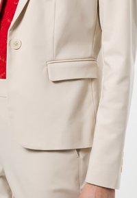 Beżowa marynarka o dopasowanym kroju, z pojedynczym guzikiem, naszywaną kieszenią i subtelną fakturą. Czerwone koronki pod spodem dodają kontrastu.