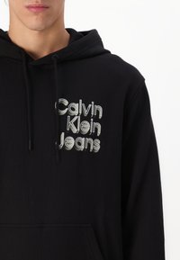 Svart bomullströja med dragsko, med en broderad "Calvin Klein Jeans"-logotyp i vitt och grått över bröstet. Framsidan har en kenguruficka.