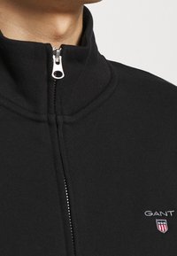 Chaqueta de forro polar negra con cremallera y cuello alto, con cremallera plateada y logo bordado de GANT en el pecho izquierdo. Textura suave.