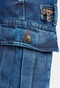 Salopette en denim avec un effet délavé bleu, fermeture éclair argentée, détails en bouton métallique et surpiqûres rouges. Le design plissé ajoute de la texture.