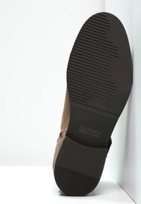 Stivaletto in suede marrone con suola in gomma nera, caratterizzato da una superficie testurizzata e un dettaglio con marchio. Tacco piatto con scanalature verticali per una migliore aderenza.